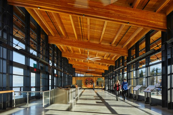 timber beams and ceiling with large windows and two people walking inside terminal building, one in blue and one in red shirt