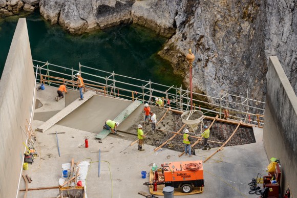 Men working in hardhats and vests, on a concrete dam spillway with river in background