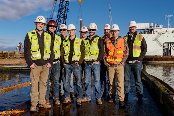 Construction men wearing high visibility vests standing at Fairhaven shipyard with crane in background and blue sky