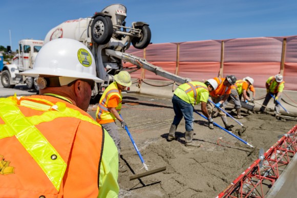 Construction workers in hard hats and high visibility vests working on I-90 roadway to place concrete during a closure, with blue sky in background