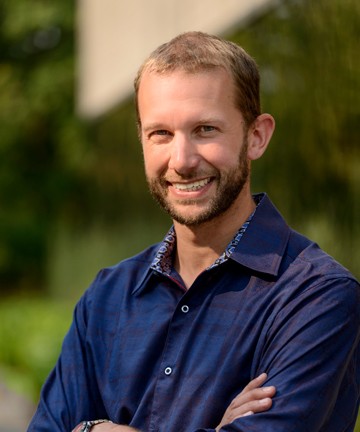 Headshot of a man standing outside smiling, in navy shirt, with arms crossed 