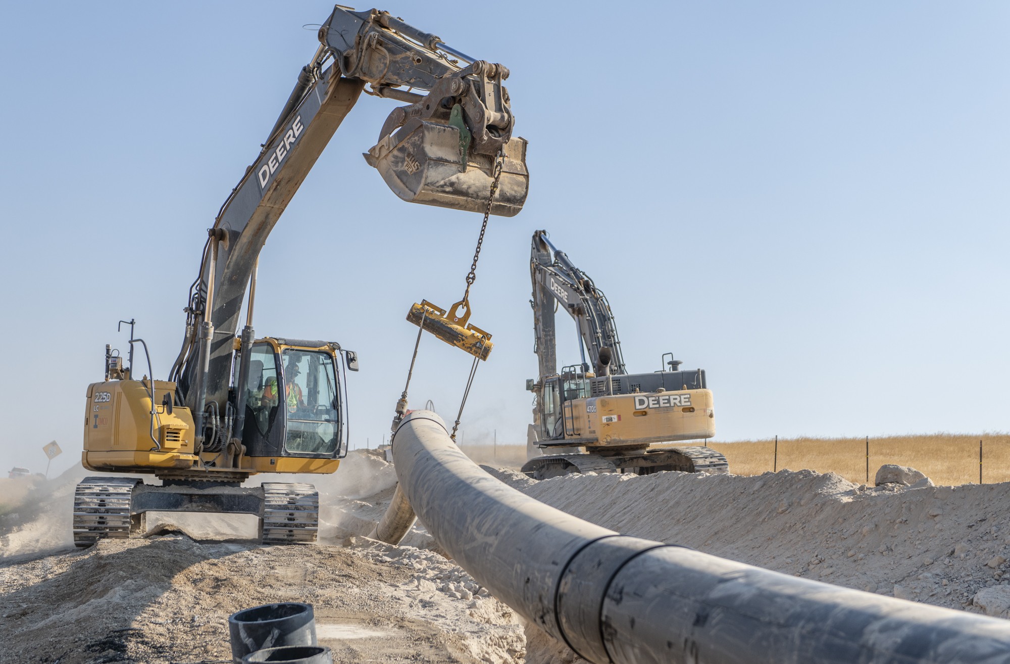 Two excavators and construction workers hoisting black pipe into a dirt trench