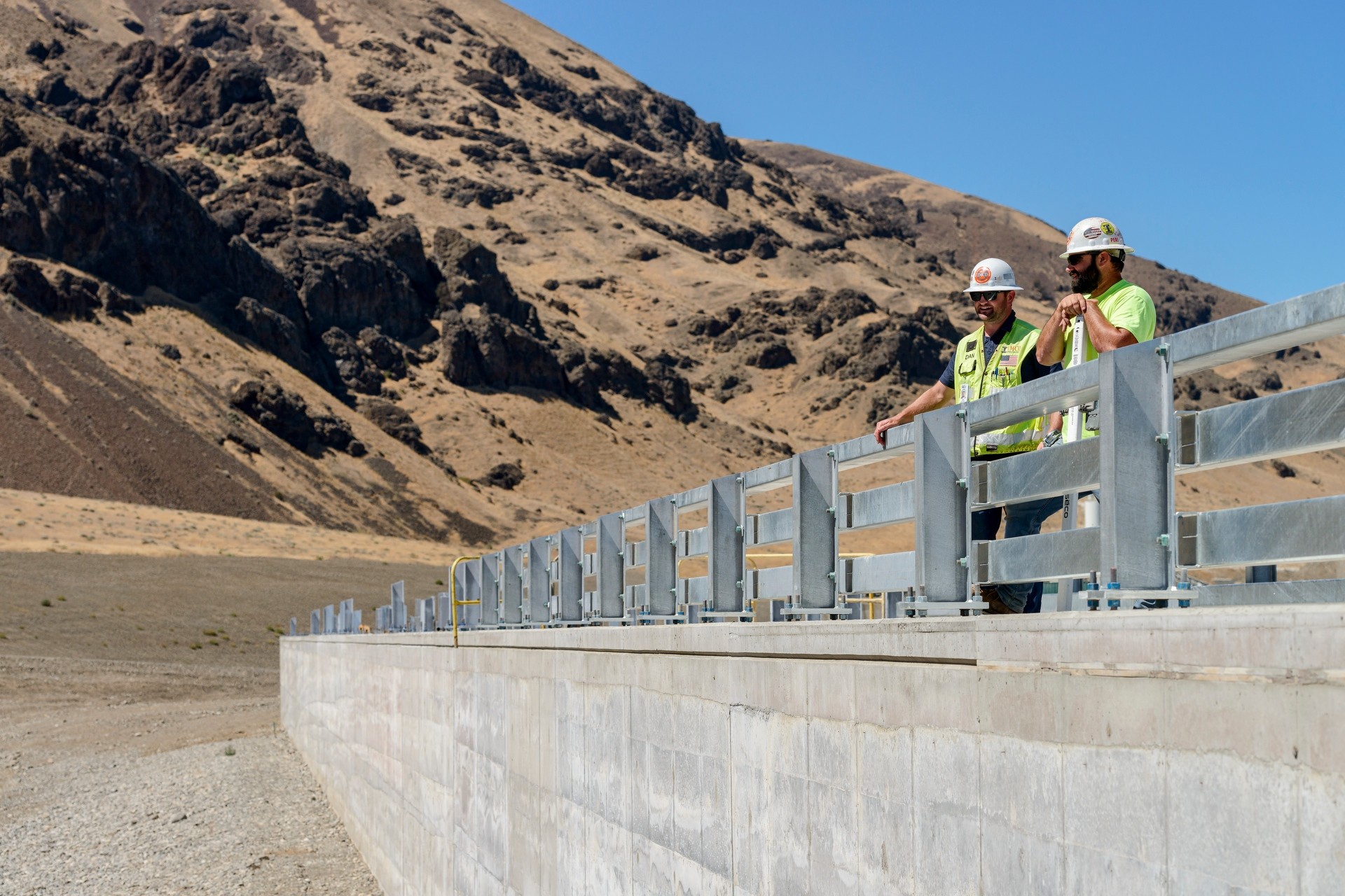 two construction workers standing on a dam with brown hills behind