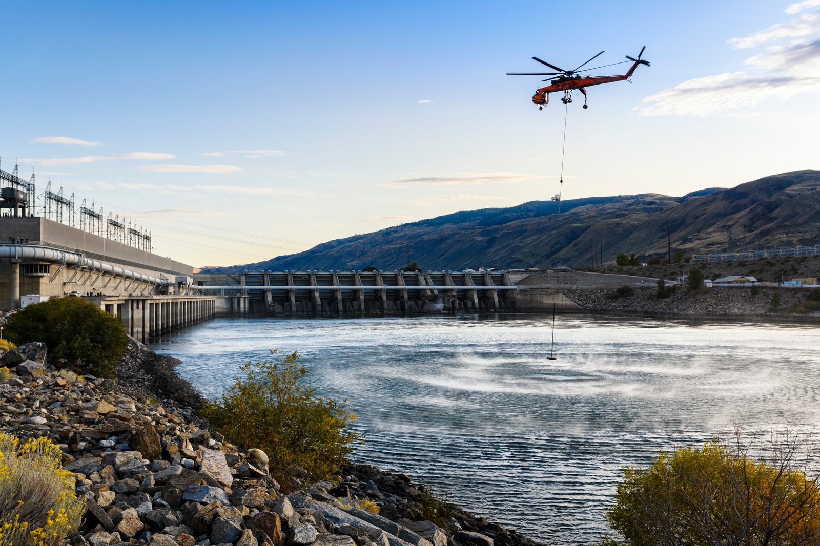 helicopter flying over river and dam facility