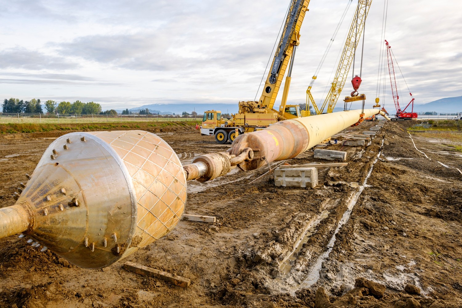 Construction site with large pipeline, red crane, yellow crane, excavator, and truck in mud
