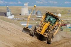 yellow grader with GPS gear and red train and industrial plant in background