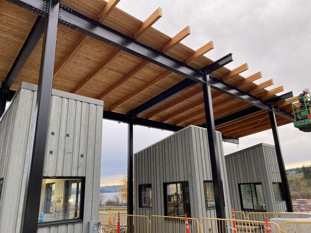 Modern building under construction with black steel framing and exposed timber roof canopy spanning over gray vertical-clad structures, as workers use a lift on an overcast day.
