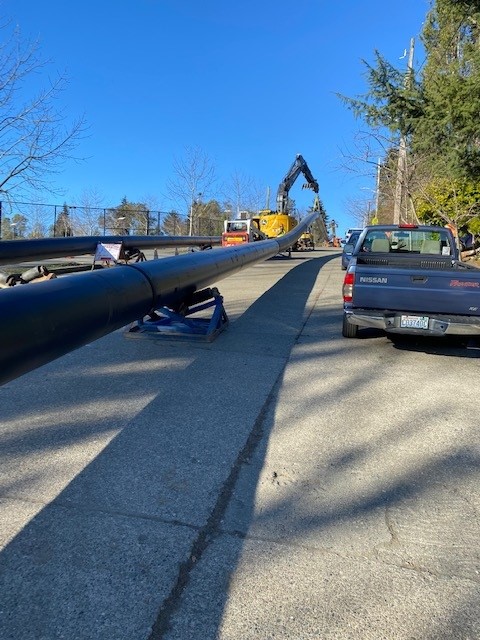 Construction crew pulling a long black pipeline along a roadside using heavy equipment, with trucks parked nearby on a clear day.