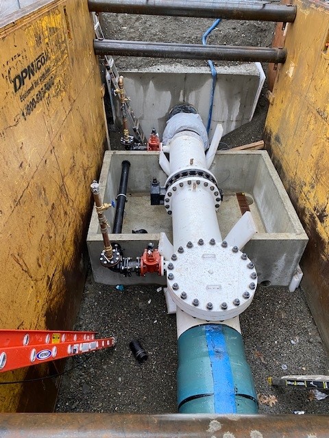 Large underground water pipeline connection inside a construction pit, featuring a bolted valve assembly, concrete supports, and surrounding excavation equipment.