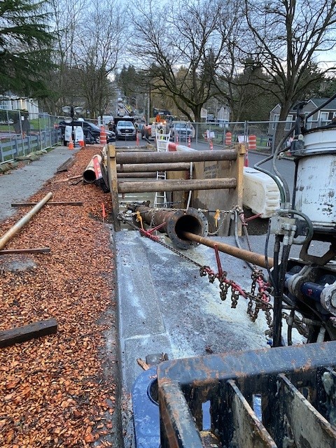 Utility crew installing underground pipe along a residential street using heavy equipment, with traffic cones, fencing, and construction vehicles lining the road.