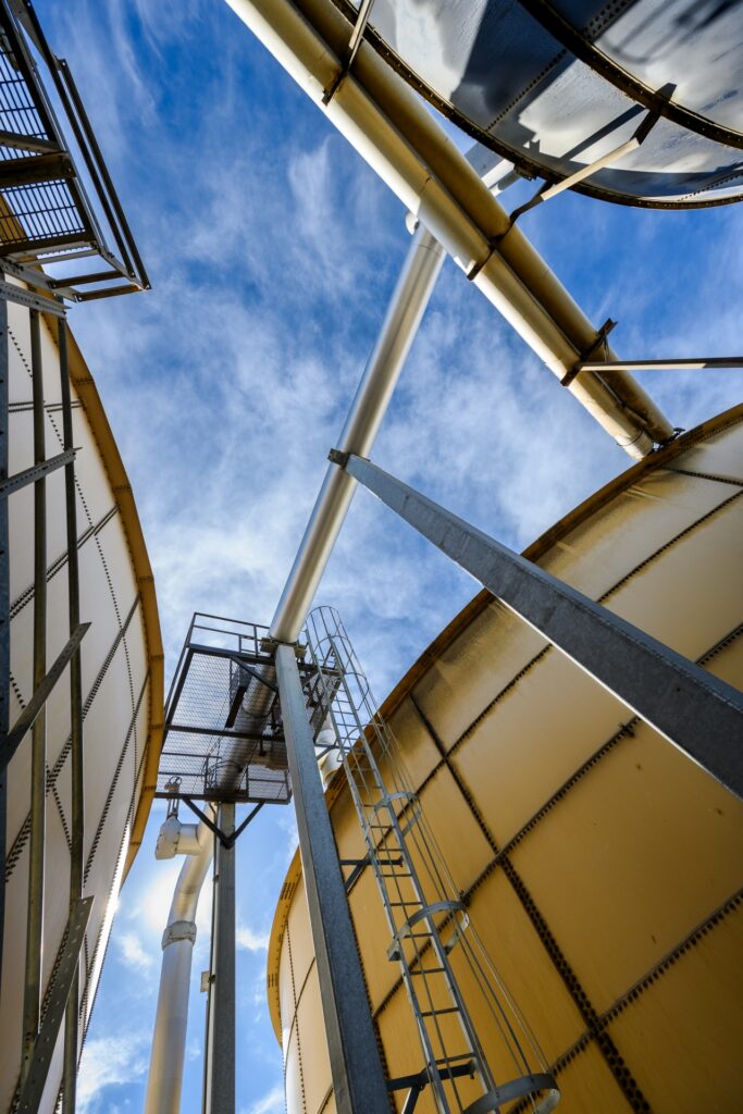Low-angle view of large industrial storage tanks with metal pipes, ladders, and platforms against a blue sky with light clouds.