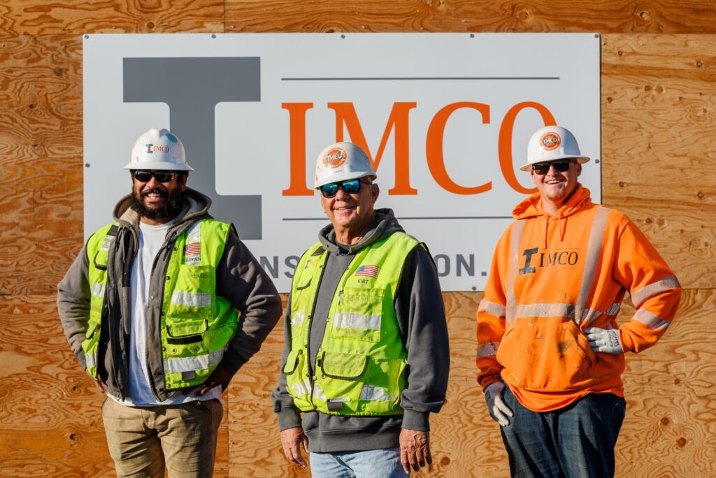 Three construction crew members standing, smiling, in front of IMCO Construction logo