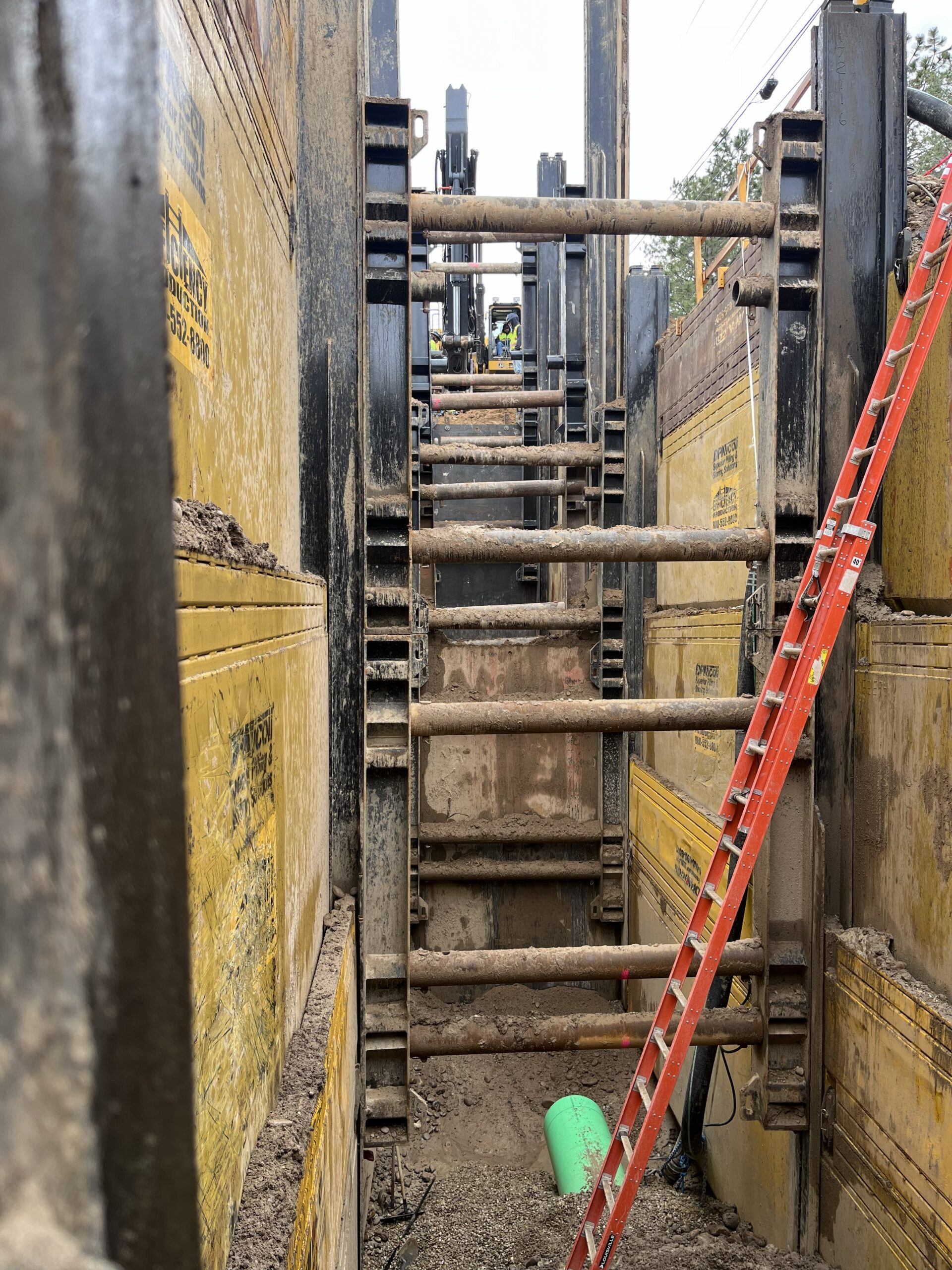 Deep construction trench reinforced with yellow steel shoring panels and cross braces, with an orange ladder leaning down into the excavation and a green pipe segment at the bottom.