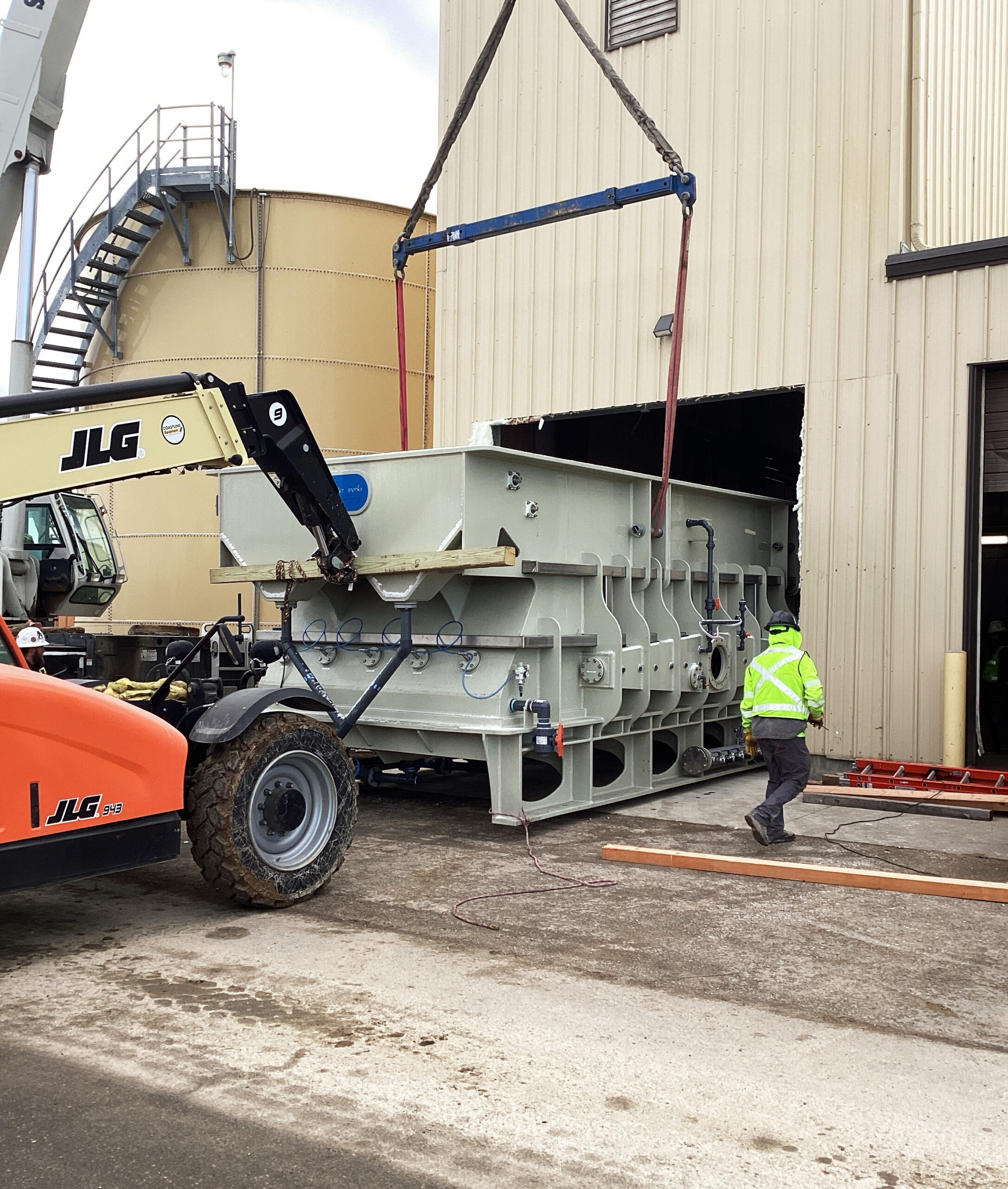 Worker in high-visibility jacket guides a crane lifting a large industrial tank with a telehandler outside a factory building.