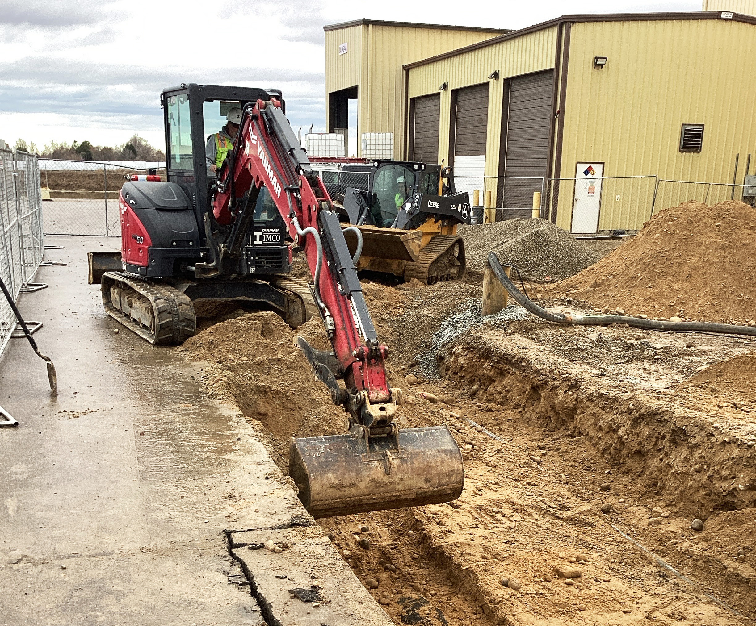 Compact excavator digging a trench beside a building at a construction site, with a skid-steer loader and piles of dirt and gravel in the background.
