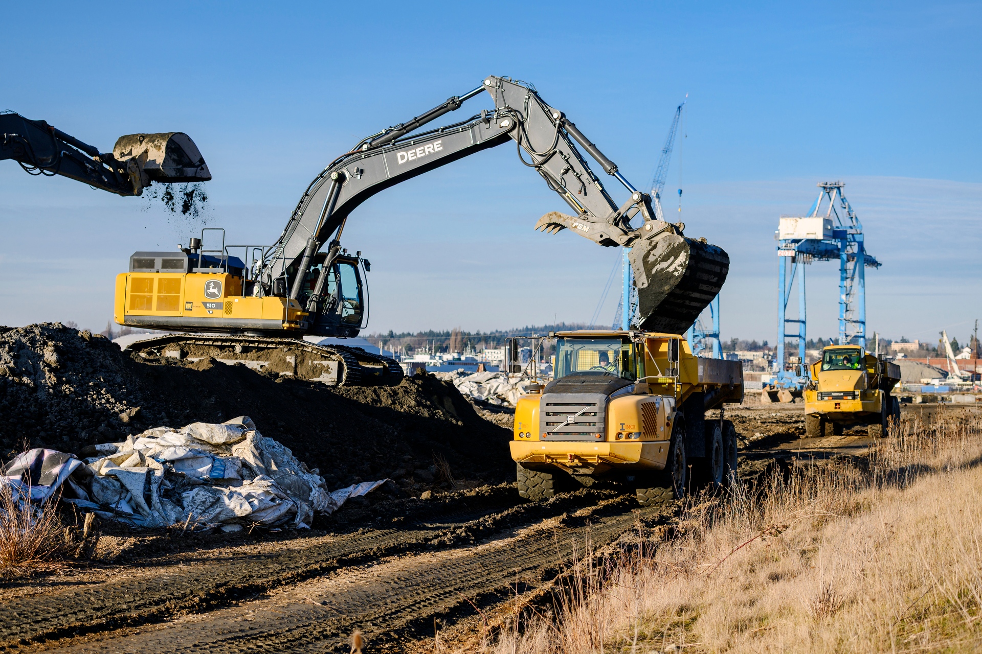 An excavator loads soil into a yellow dump truck at a construction site with large blue shipyard cranes in the background.