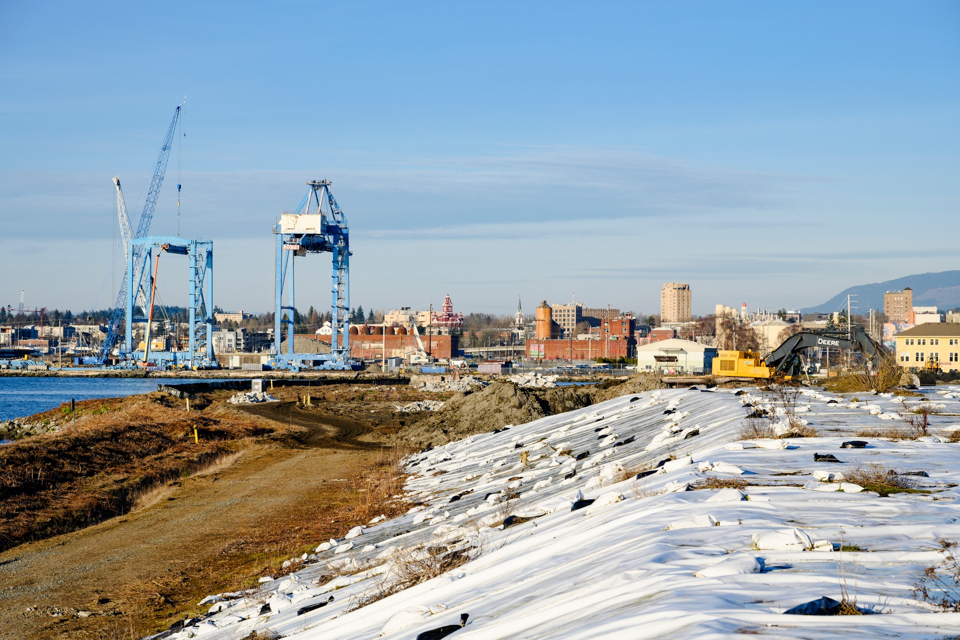 An excavator works on a shoreline construction site with large blue shipyard cranes and a city skyline in the background.