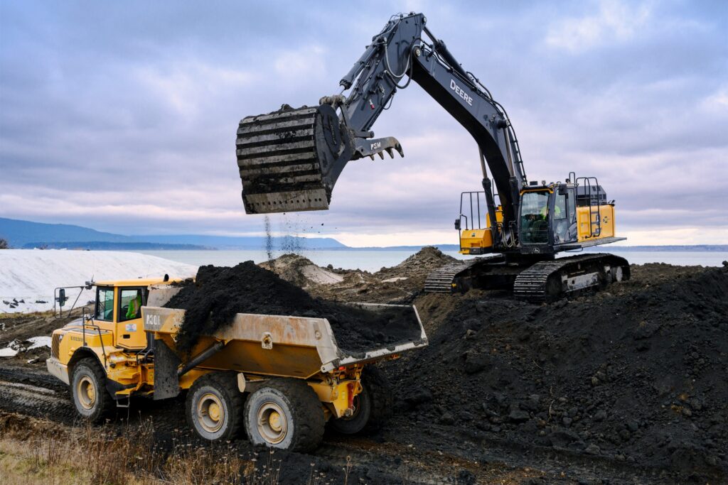 A large excavator loads dark soil into a yellow dump truck at a construction site near a body of water under a cloudy sky.