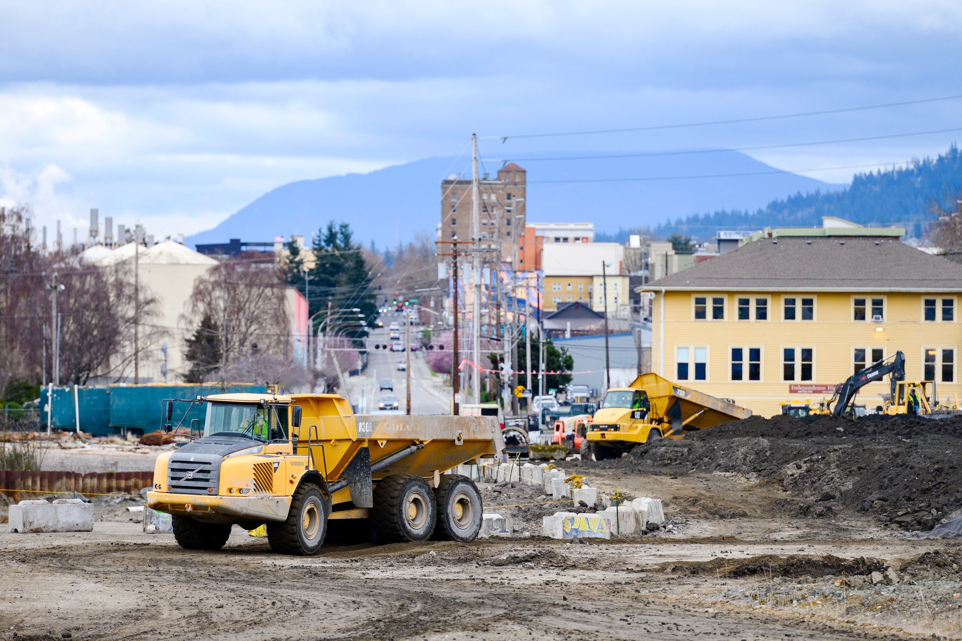 A yellow dump truck sits at a construction site with another truck and an excavator working in the background along a city street.