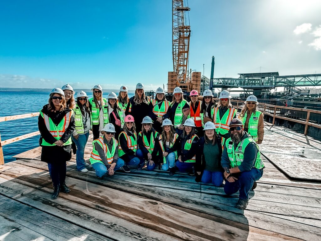 Group of women in safety gear posing on a dock over the ocean with a crane in the background.