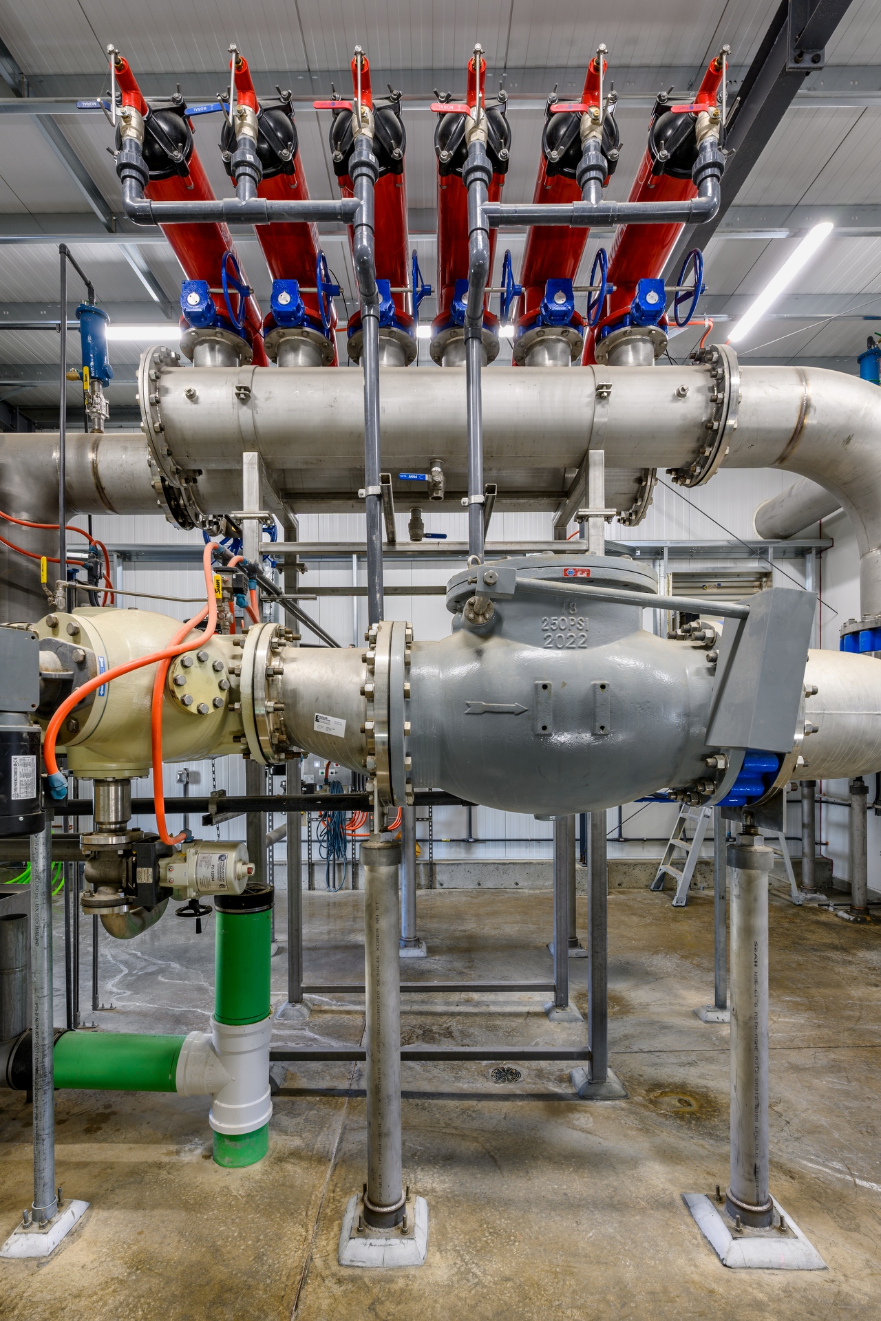 Industrial piping system inside a facility, featuring a large central valve assembly, stainless steel pipes, red vertical filter housings, blue handwheel valves, and control lines mounted on a concrete floor.