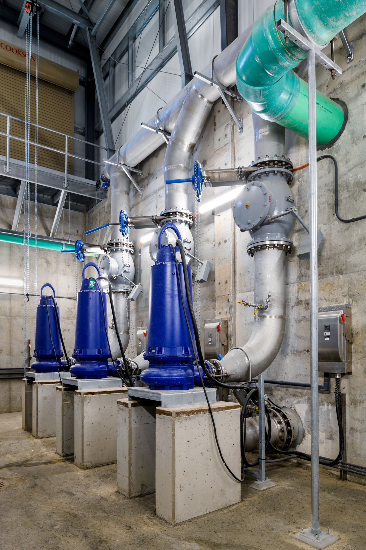Interior of an industrial pump room with four large blue pumps mounted on concrete bases, connected to overhead metal piping, valves, and control panels along a concrete wall.