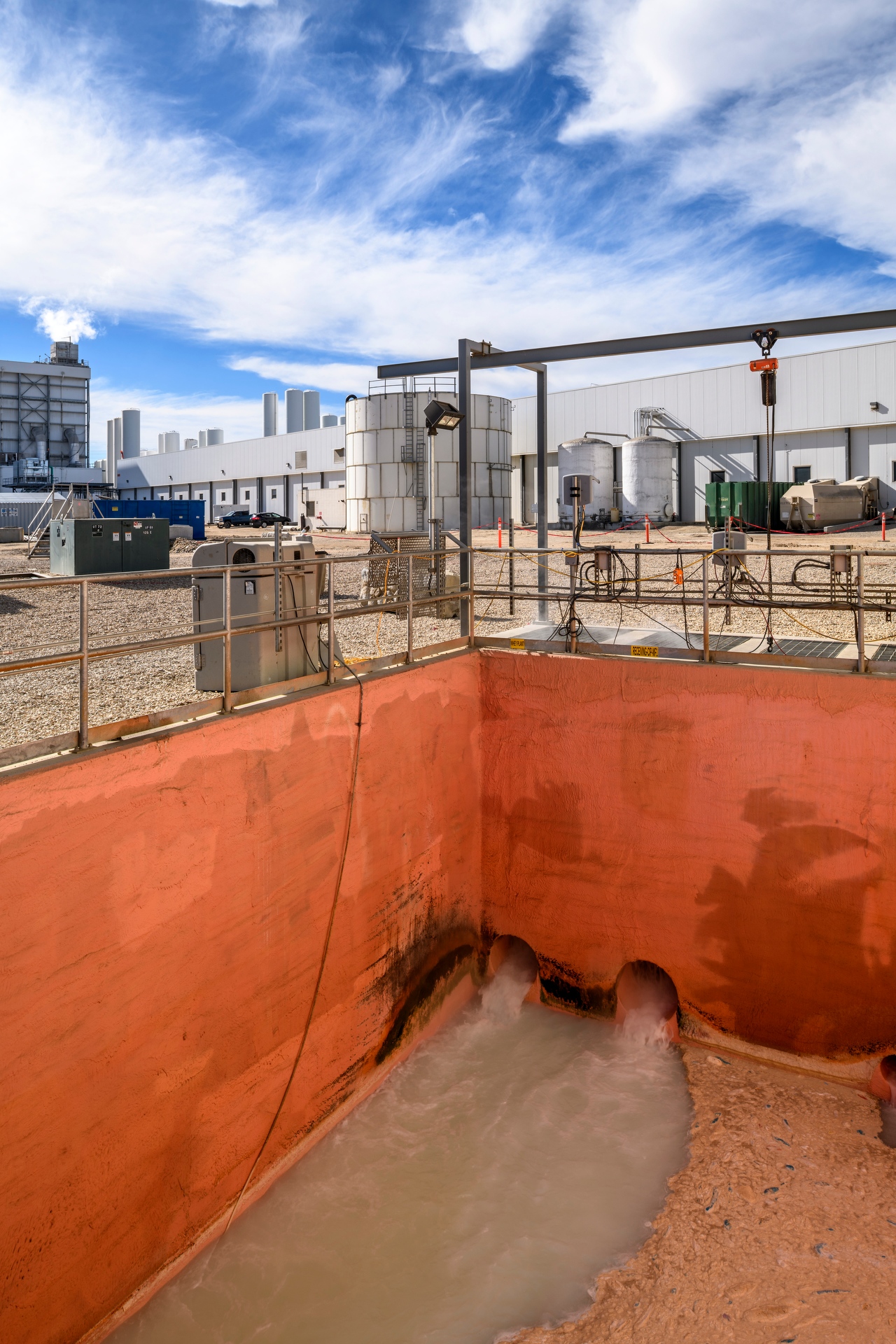 Outdoor industrial wastewater pit with orange concrete walls, where cloudy water flows from two large pipes, surrounded by railings and equipment at a processing facility under a blue sky.