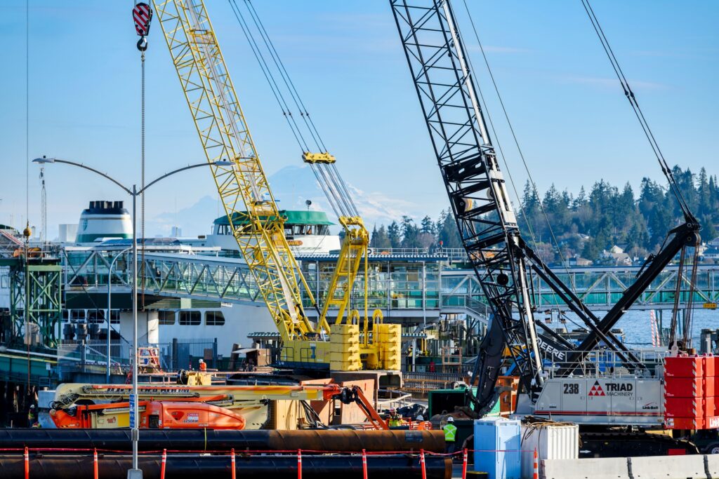 Construction activity at an active ferry terminal, featuring multiple cranes lifting materials near a docked ferry, with workers on site and Mount Rainier visible in the background.