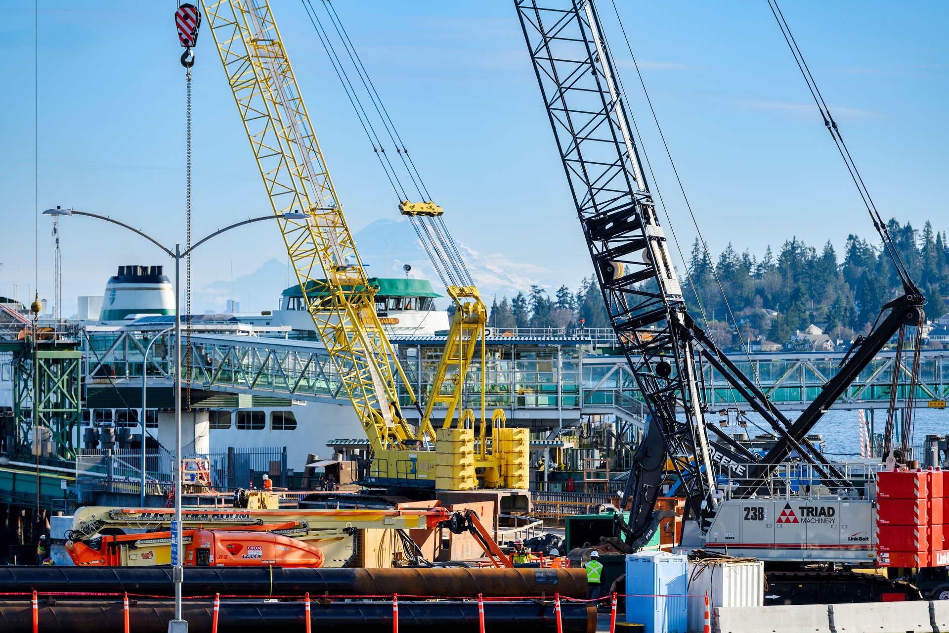 Construction activity at an active ferry terminal, featuring multiple cranes lifting materials near a docked ferry, with workers on site and Mount Rainier visible in the background.
