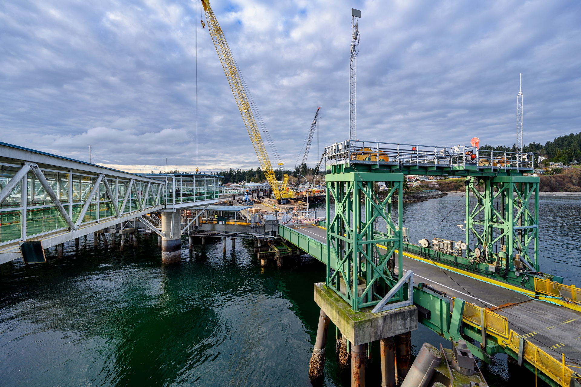 Wide view of an active ferry terminal construction site, showing cranes, elevated walkways, and marine structures extending over the water.