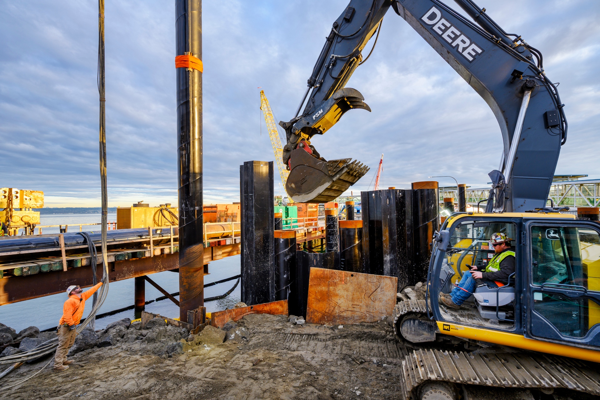 Excavator placing material around steel piles at a waterfront construction site while a worker guides positioning near an active ferry terminal.