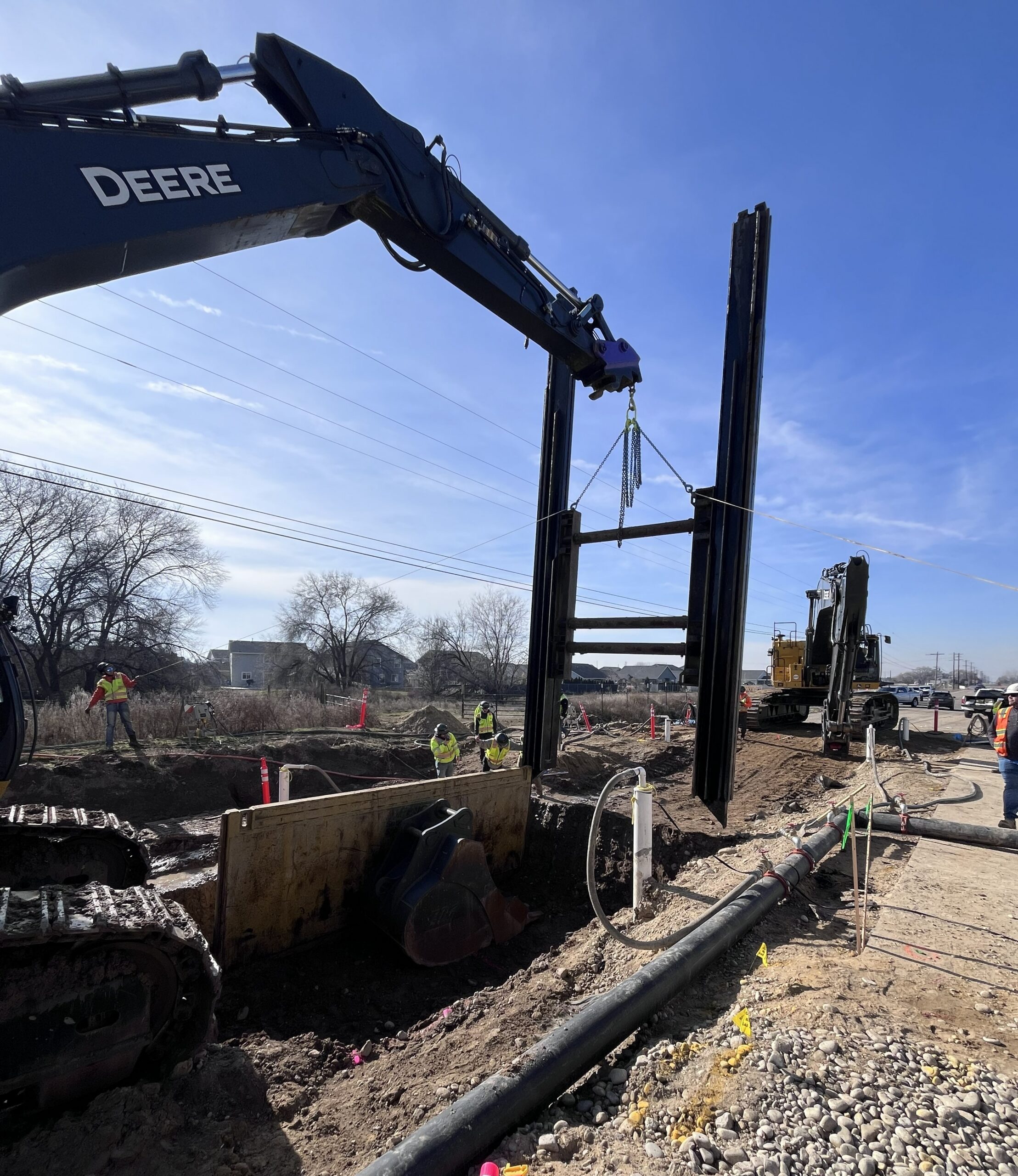 Construction crew installing large steel trench shoring panels with an excavator at a roadside excavation site under a clear blue sky. Workers in high-visibility safety gear stand around the trench while heavy equipment operates nearby.