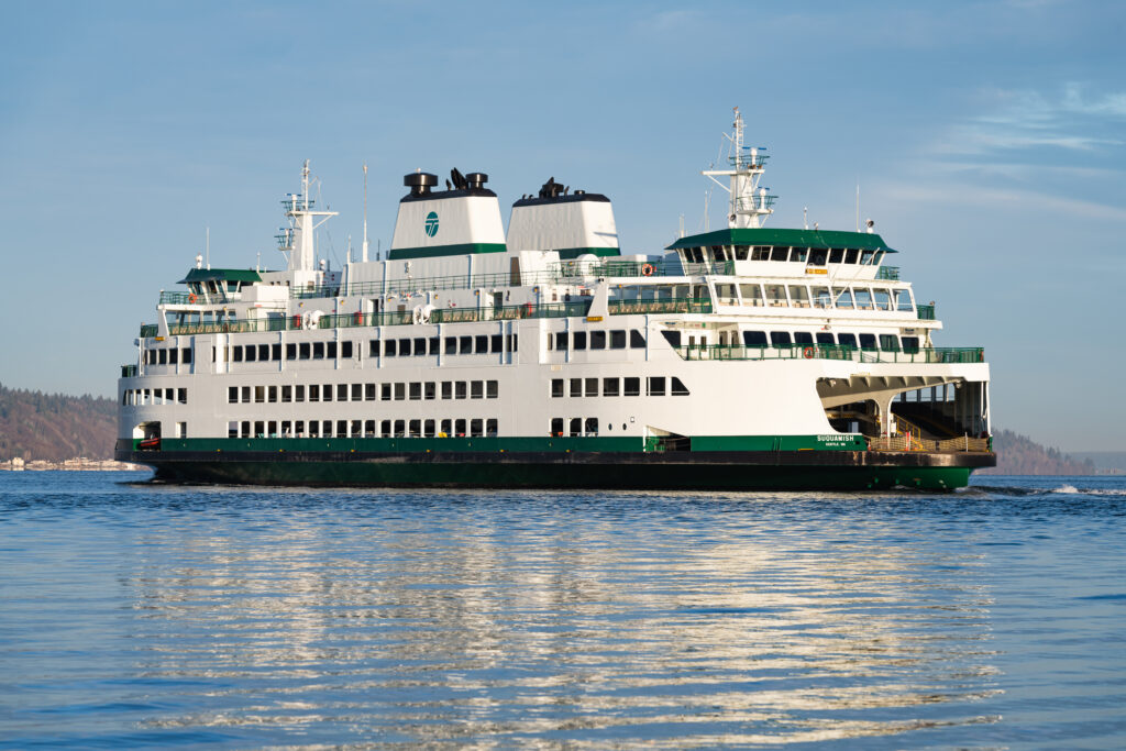 A large white-and-green passenger ferry travels across calm blue water under a partly cloudy sky, with vehicle loading ramps visible at both ends and shoreline trees in the distance.