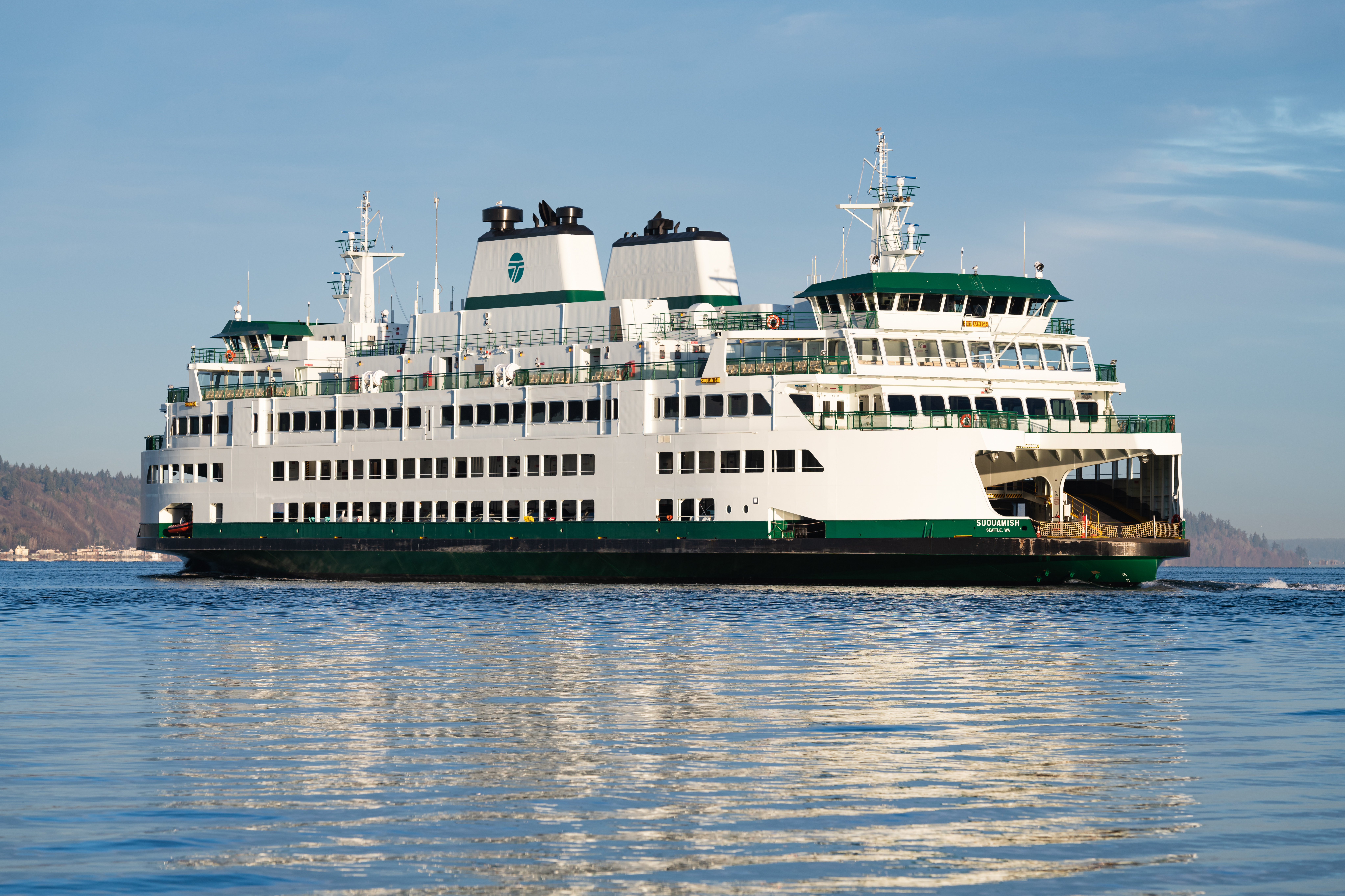 A large white-and-green passenger ferry travels across calm blue water under a partly cloudy sky, with vehicle loading ramps visible at both ends and shoreline trees in the distance.