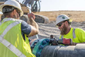 Two construction workers in hard hats and high-visibility safety vests install a large pipeline flange at an outdoor jobsite. One worker tightens a large wrench while the other operates a powered torque wrench on the flange bolts, with an excavator visible in the background.