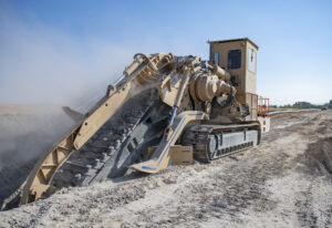 A tracked trenching machine excavates a deep trench at a pipeline construction site, cutting through dry soil and creating a cloud of dust. The machine operates along a dirt access road under a clear blue sky, with the pipeline corridor extending into the distance.