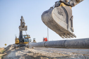 Close-up of an excavator lowering a large bucket near a newly installed black pipeline at an active construction site. Additional heavy equipment is visible in the background as the pipeline extends across a graded dirt trench under a clear blue sky.