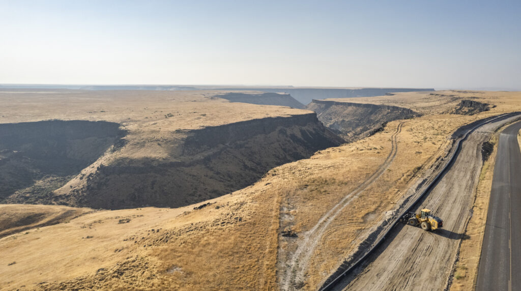 A wide view of a dry canyon landscape with golden grass-covered cliffs stretching into the distance under a clear blue sky. On the right, a road runs alongside an active construction area where a large loader is working on a newly graded roadway near the canyon's edge.