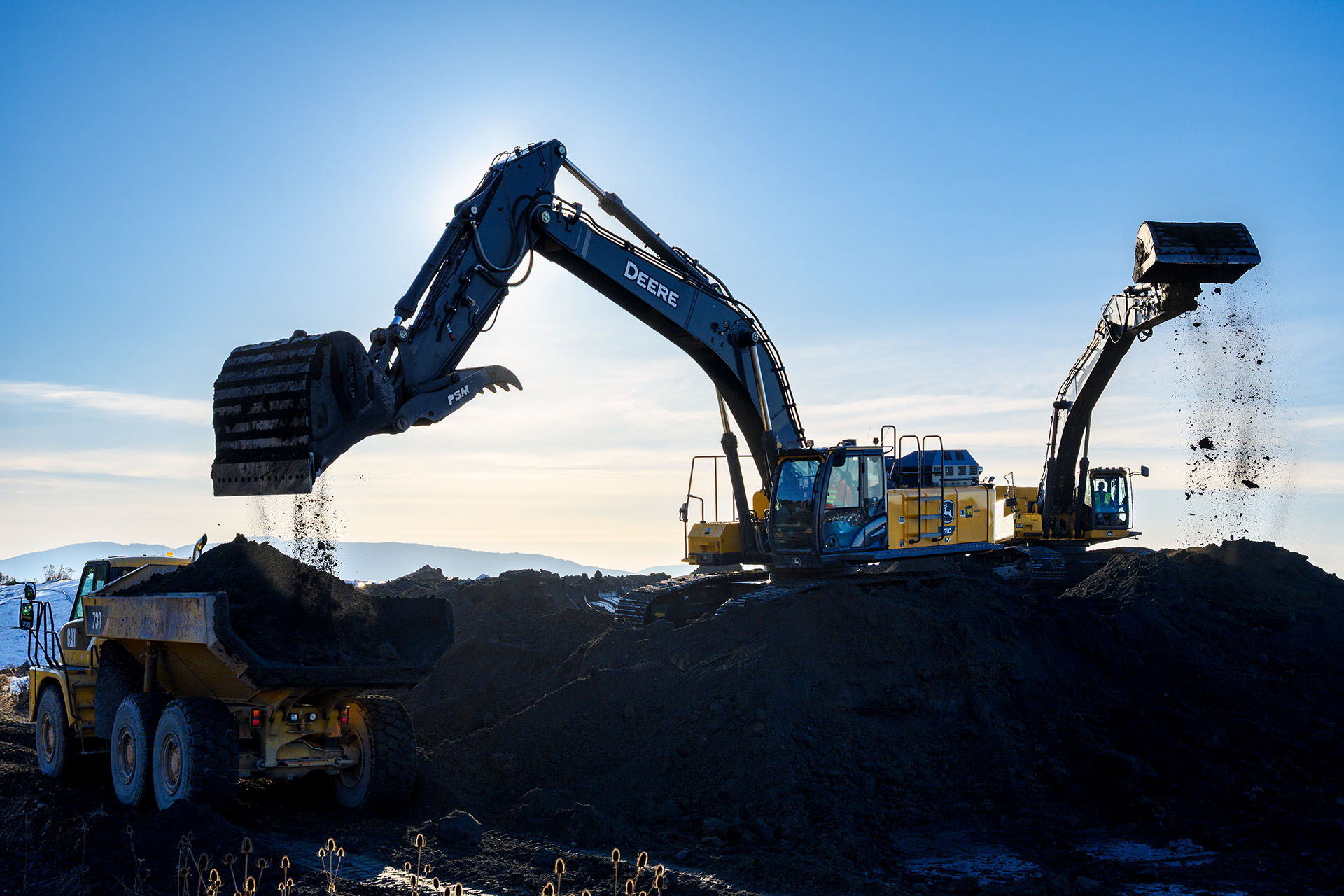 backlit excavators moving dirt along coastline with blue sky in background