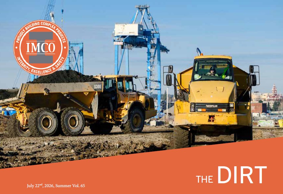 Two yellow articulated dump trucks operate on a large earthwork construction site. One truck, loaded with soil, is positioned on the left while another faces the camera on a dirt haul road with its operator visible inside the cab. Large blue shipyard cranes rise in the background under a clear blue sky, highlighting the site's proximity to a waterfront industrial area. Freshly graded ground and heavy equipment tracks fill the foreground.